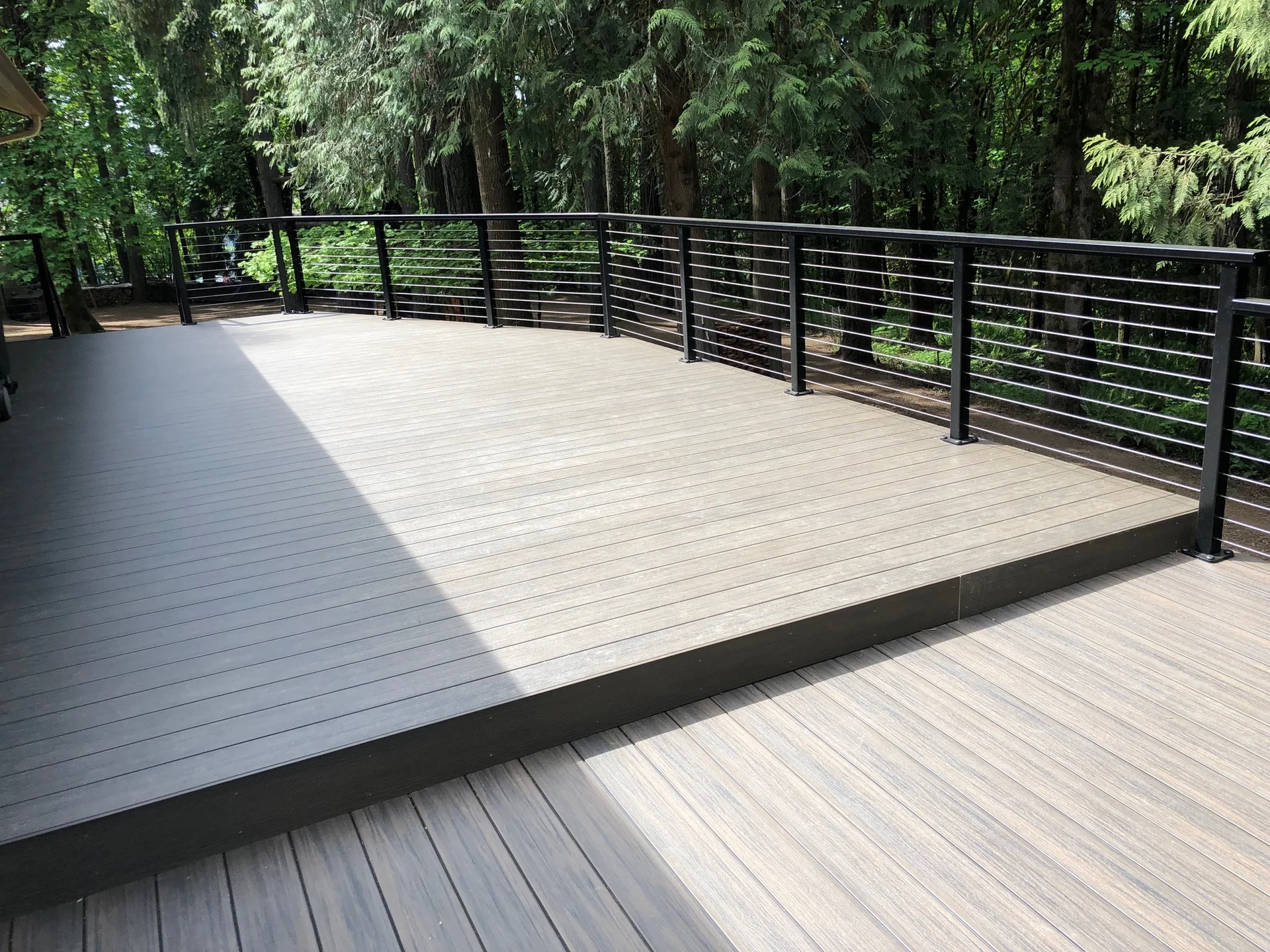 Two-level gray composite deck edged with black cable railing and mature trees